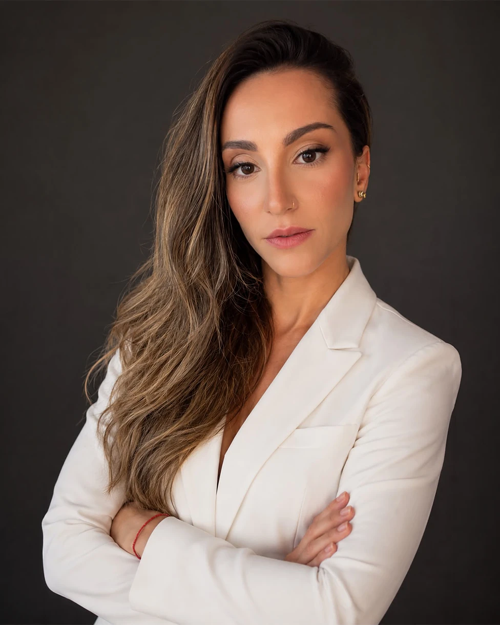 Dra. Thaís Fonseca, retrato com fundo escuro, blazer branco e braços cruzados